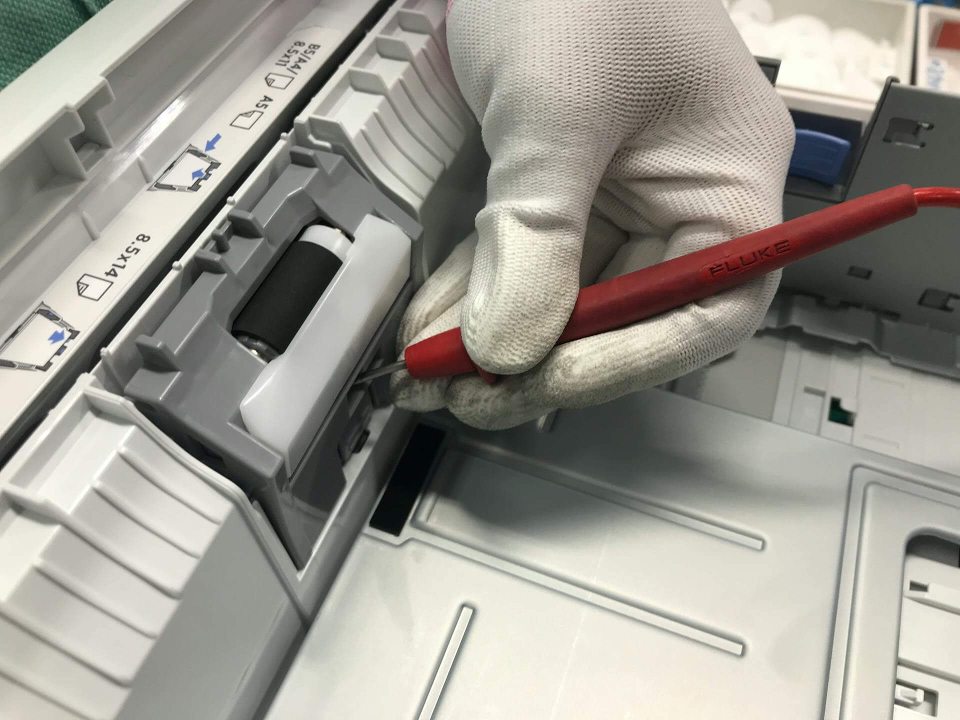 Technician repairing printer mechanism with Fluke tool. Printer paper size indicators visible.